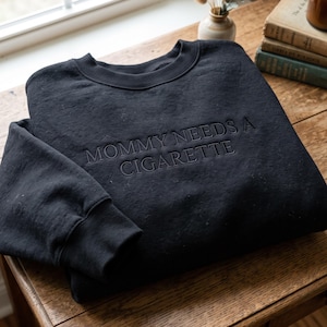 May include: A black crewneck sweatshirt featuring the embroidered text MOMMY NEEDS A CIGARETTE in a matching black thread. The garment has long sleeves with ribbed cuffs and a ribbed neckline, showcasing a monochromatic design on the front chest area.