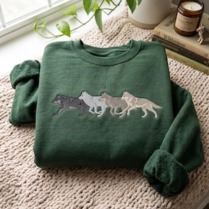 Puede incluir: Una sudadera de cuello redondo en color verde bosque oscuro con un gráfico bordado de cuatro lobos corriendo en fila. Los lobos están representados en varios tonos de gris, blanco y beige, dispuestos horizontalmente en el centro del pecho.