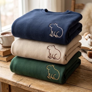 Op de afbeelding: Een stapel van drie sweaters met ronde hals in marineblauw, beige en donkergroen. Elke sweater is voorzien van een eenvoudig, geborduurd lijnontwerp van een capibara op de borst, wat zorgt voor een strakke en eenvoudige uitstraling.