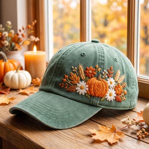 May include: A sage green baseball cap featuring detailed embroidery of a central orange pumpkin surrounded by white daisies, orange flowers, and wheat stalks. The cap has a curved brim and stitched eyelets on the crown, showcasing a textured fabric.