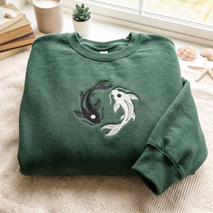 Könnte beinhalten: Ein waldgrüner Rundhals-Pullover mit einer Stickerei von zwei Koi-Fischen, einem schwarzen und einem weißen, die in einem kreisförmigen Muster angeordnet sind. Die Fische sind so positioniert, dass sie ein Yin-und-Yang-Symbol auf der Brust bilden.