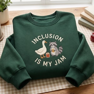 Op de afbeelding: Een donkergroene sweater met ronde hals en een borduursel van een witte gans en een grijze wasbeer die een pot jam vasthoudt. De tekst "INCLUSION IS MY JAM" is in het wit boven en onder de dieren geborduurd.