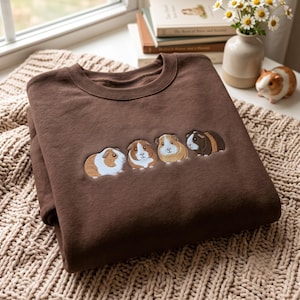 Op de afbeelding: Een donkerbruin sweatshirt met ronde hals en een geborduurd ontwerp van vier cavia's op een rij. De cavia's zijn afgebeeld in verschillende kleurpatronen, waaronder wit, bruin en donkerbruin, met gedetailleerde stiksels en texturen.