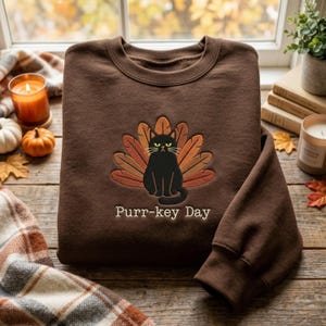 Peut inclure: Un sweat-shirt à col rond marron foncé avec une broderie représentant un chat noir assis devant un éventail de plumes orange, marron et beige. Sous le chat, le texte Purr-key Day est brodé dans une police blanche simple.