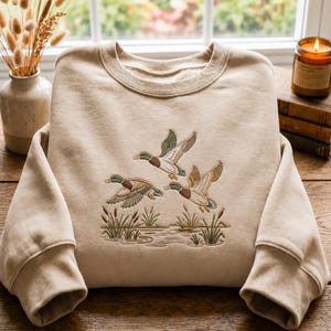 Puede incluir: Una sudadera beige de cuello redondo con un diseño bordado de tres patos ánades reales volando sobre un paisaje pantanoso con juncos y agua. El bordado utiliza tonos verdes, marrones, blancos y beige para representar los patos y plantas.