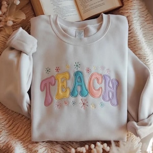 Könnte beinhalten: Ein hellbeiges Sweatshirt mit Rundhalsausschnitt, auf dem das Wort TEACH in großen, bunten Buchstaben im Bubble-Stil gestickt ist. Jeder Buchstabe hat einen anderen Pastellton, darunter Rosa, Gelb, Blau, Orange und Lila, ergänzt durch kleine Sternmotive.