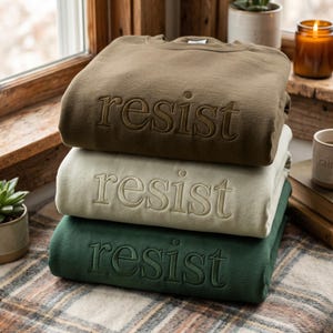 May include: A stack of three crewneck sweatshirts in olive green, cream, and dark forest green. Each sweatshirt features the word "resist" embroidered in a matching thread colour across the centre chest in a classic serif font style.