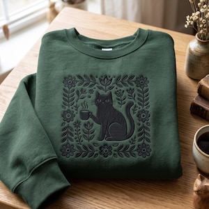May include: A forest green crewneck sweatshirt featuring a dark embroidered graphic of a cat holding a steaming mug, centred within a square frame of stylised flowers and leaves. The embroidery is tonal, matching the dark green fabric.