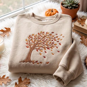 Pode incluir: Uma camisola de gola redonda bege com um design bordado de uma árvore com ramos soprados pelo vento e folhas de outono a cair em tons de laranja, vermelho e castanho. O bordado está centrado no peito da peça de manga comprida.