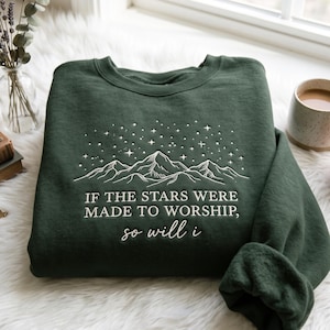 Könnte beinhalten: Ein dunkelwaldgrüner Rundhals-Pullover mit weißer Stickerei eines Gebirges unter einem Sternenhimmel. Der Text "IF THE STARS WERE MADE TO WORSHIP, so will i" ist in weiß unter der Berggrafik eingestickt.