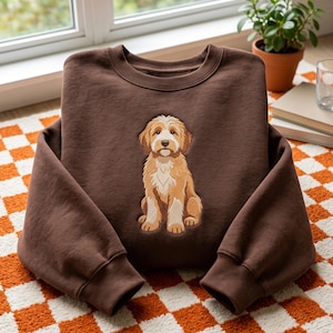 Könnte beinhalten: Ein dunkelbraunes Sweatshirt mit Rundhalsausschnitt und einer mittigen grafischen Illustration eines sitzenden, zotteligen Hundes in Hellbraun und Weiß. Der Hund hat Schlappohren und einen weißen Fleck auf der Brust.