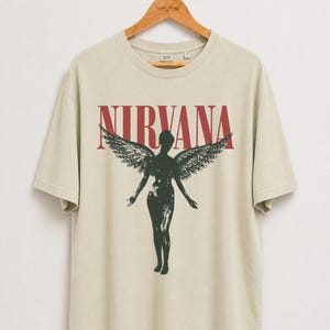 Peut inclure: Un t-shirt beige à manches courtes avec le mot NIRVANA en lettres capitales rouges au-dessus d'un graphique noir représentant une silhouette ailée. Le t-shirt possède un col rond et est présenté sur un cintre en bois.
