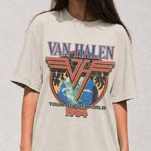 May include: A beige short-sleeve t-shirt featuring a graphic print of the Van Halen logo above an illustration of the Earth. The text reads VAN HALEN, TOUR OF THE WORLD, and 1984 in a distressed, multi-coloured font with red, blue, and yellow accents.