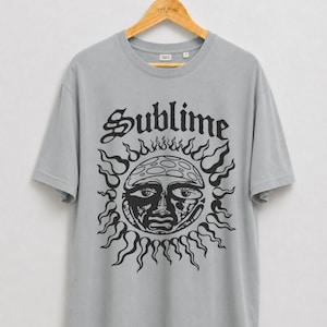 Puede incluir: Una camiseta gris claro de manga corta con un estampado gráfico negro de un sol con rostro humano y la palabra Sublime en una fuente gótica encima. La camiseta tiene cuello redondo y se muestra en una percha de madera.
