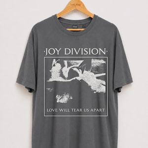 Puede incluir: Una camiseta de manga corta en color gris carbón con un estampado gráfico en blanco. El diseño incluye el texto JOY DIVISION en la parte superior y LOVE WILL TEAR US APART en la inferior, enmarcando una ilustración abstracta blanca.