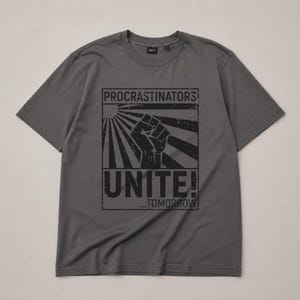 Op de afbeelding: Een antracietgrijs T-shirt met korte mouwen en een zwarte grafische print van een opgeheven vuist tegen een achtergrond van zonnestralen. De tekst luidt PROCRASTINATORS UNITE! ...TOMORROW in een verweerd bloklettertype binnen een kader.