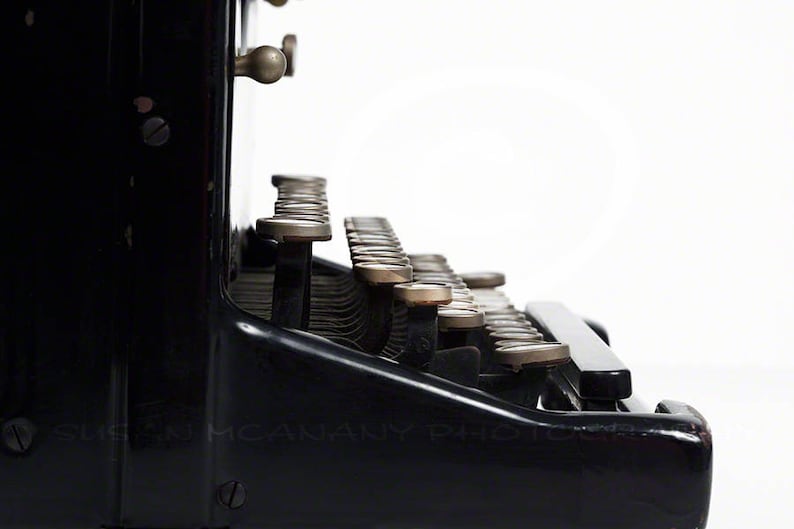 May include: A black vintage typewriter with a row of keys and a metal carriage return lever. The keys are made of metal and have a gold finish.