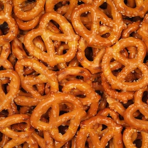 May include: A close-up image of a pile of salted pretzels. The pretzels are a light brown colour and are covered in salt crystals.
