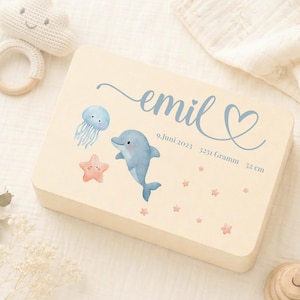 Könnte beinhalten: Eine hellfarbene Holzkiste mit einem Aquarelldesign mit einem blauen Delfin, einer Qualle und einem Seestern. Der Name "emil" ist in blauer Schrift geschrieben, zusammen mit dem Datum, dem Gewicht (3251 Gramm) und der Größe (52 cm). Ein Holzspielzeug und eine Häkelwolke befinden sich im Hintergrund.