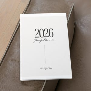 Könnte beinhalten: Ein weißer Jahresplaner 2026 mit schwarzem Text, der auf einer braunen Lederoberfläche liegt. Das Cover zeigt das Jahr 2026 in einer fetten Schrift, darunter "Yearly Planner" in einer Schreibschrift. Der Planer hat ein schlichtes, elegantes Design.