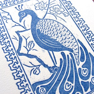 May include: A blue and white print of a peacock with its tail feathers spread out. The peacock is perched on a branch with a flower behind it. The print is framed with a decorative border.