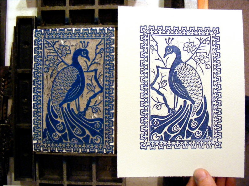 May include: A blue and white print of a peacock with its tail feathers spread out. The print is framed with a decorative border.