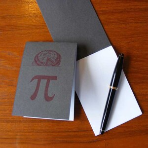 May include: A small gray notebook with a red pi symbol and a white paper pad with a black pen on a wooden surface.