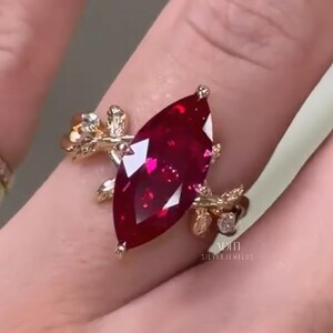 Op de afbeelding: Een gouden ring met een grote, dieprode edelsteen in marquise-slijpvorm als middelpunt. De ringband is ontworpen met gedetailleerde metalen bladmotieven en kleine ingebedde heldere steentjes. De tekst ADITI SILVER JEWELUS is zichtbaar.