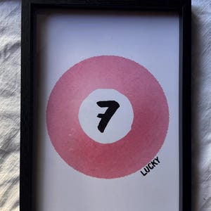 Op de afbeelding: Een ingelijste print met een roze cirkel in aquarel-stijl op een witte achtergrond. In het midden van de cirkel bevindt zich een wit vlak met het zwarte cijfer 7. Het woord LUCKY staat in zwarte hoofdletters in de rechterbenedenhoek gedrukt.