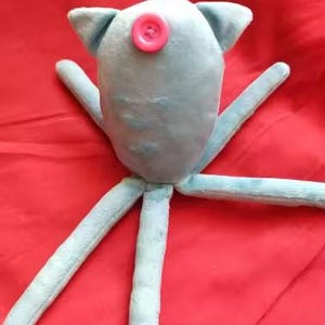 May include: A light blue plush toy with a pink button eye and four long, slender arms. The toy has small, triangular ears and is set against a red background. The soft toy is likely handmade.