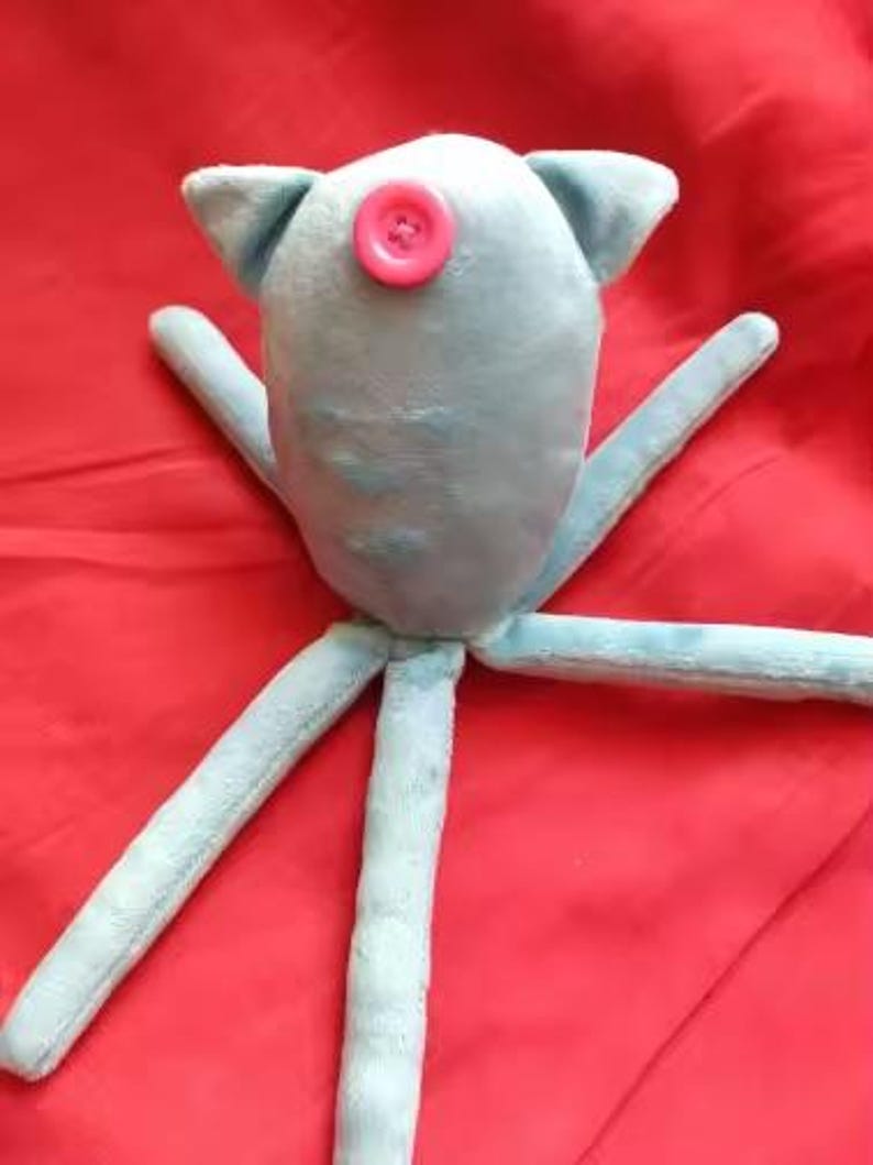 May include: A light blue plush toy with a pink button eye and four long, slender arms. The toy has small, triangular ears and is set against a red background. The soft toy is likely handmade.