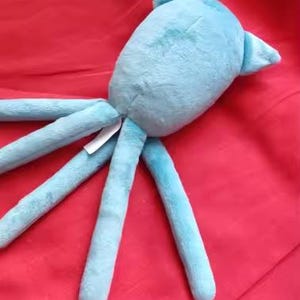 May include: A light blue plush octopus toy with long tentacles and a rounded body. The toy is made of soft material and is lying on a red surface. A small tag is attached to the toy.