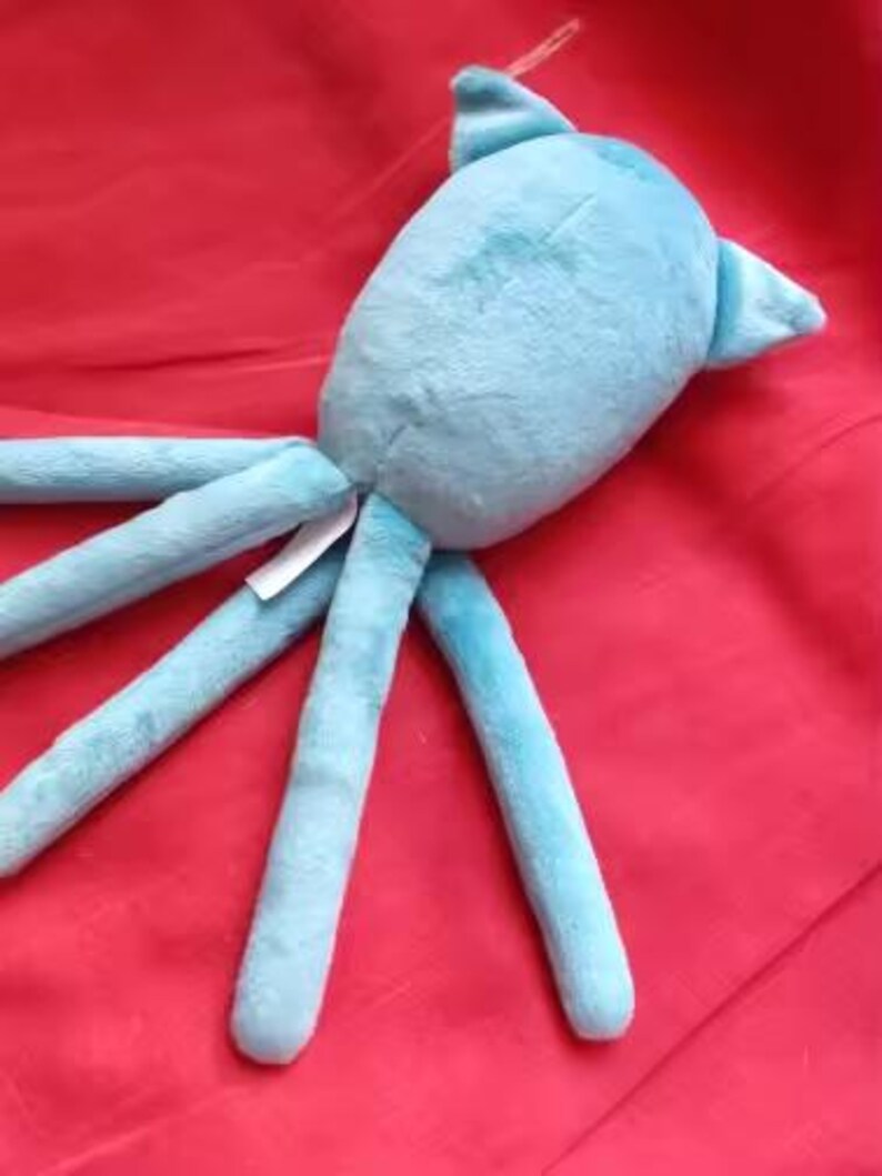 May include: A light blue plush octopus toy with long tentacles and a rounded body. The toy is made of soft material and is lying on a red surface. A small tag is attached to the toy.