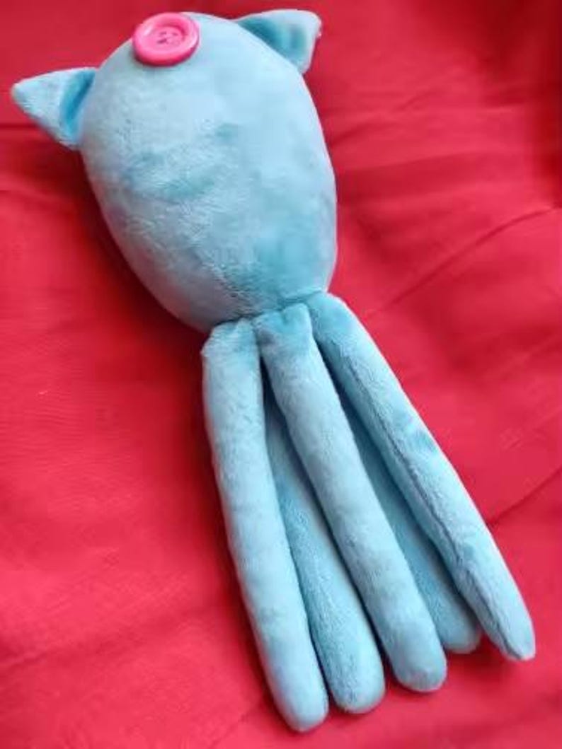 May include: A light blue plush octopus toy with a pink button on its head. The toy has two small ears and eight long tentacles. The toy is made of soft material and is lying on a red surface.