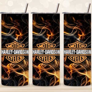 Op de afbeelding: Drie cilindrische roestvrijstalen bekers met deksels en metalen rietjes, met een zwarte achtergrond met oranje en gele rookpatronen en het oranje Harley-Davidson Motor Cycles logo horizontaal in het midden gedrukt.