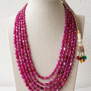 May include: A multi-strand necklace featuring five rows of polished, oval-shaped magenta beads. The necklace includes an adjustable cord closure with a decorative tassel end adorned with small, colorful beads in red, green, and yellow hues.