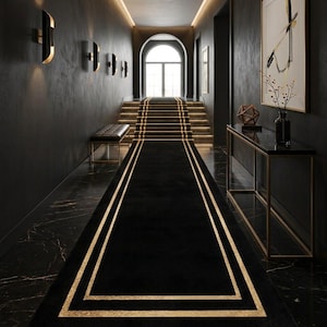 May include: A long rectangular black runner rug featuring two concentric gold-colored rectangular borders. The rug is positioned centrally on a dark marble floor, extending from the foreground toward a set of stairs in a hallway with dark walls.