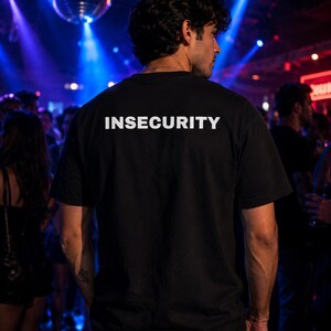 以下が含まれることがあります： 背面上部に太字の白い大文字で「INSECURITY」という単語がプリントされた黒の半袖Tシャツ。この衣服はクラシックなクルーネックと標準的なフィット感を備えており、暗い場所で背面から見た様子です。