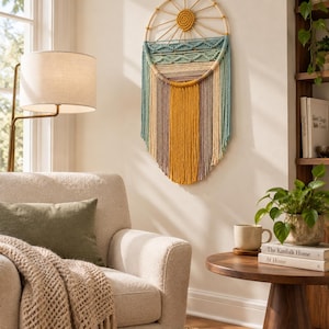 May include: A circular macrame wall hanging featuring a central sun motif made of mustard yellow cord. Below the sun, layers of knotted cord in shades of teal, cream, and grey form a landscape pattern, with long, vertical fringe in yellow and grey.