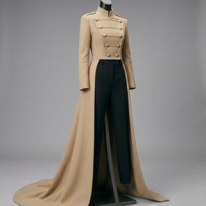 Op de afbeelding: Een beige jas in militaire stijl met gouden knopen en een hoge kraag, gecombineerd met een zwarte broek. De jas heeft een lang, vloeiend ontwerp, gepresenteerd op een mannequin tegen een grijze achtergrond. De jas is ongeveer 152 cm lang.