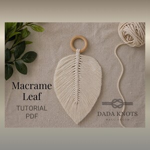May include: A cream macrame leaf wall hanging with a wooden ring and central vein design. The image includes a ball of yarn and the text "Macrame Leaf TUTORIAL PDF" and "DADA KNOTS WALL DECOR".