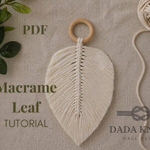 May include: A cream-coloured macrame leaf with a wooden ring at the top. The leaf is made of woven cotton cord and has a detailed, textured design. The image also includes a ball of cord and the text "Macrame Leaf Tutorial".