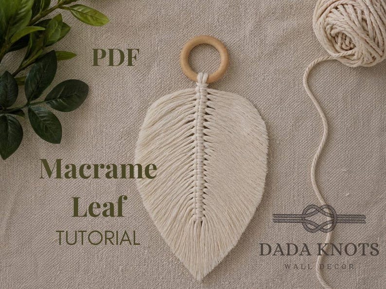 May include: A cream-coloured macrame leaf with a wooden ring at the top. The leaf is made of woven cotton cord and has a detailed, textured design. The image also includes a ball of cord and the text "Macrame Leaf Tutorial".