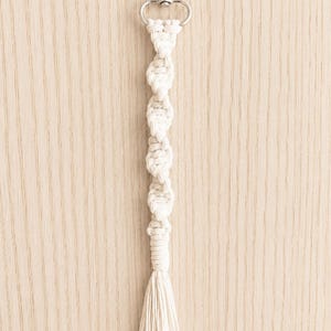 Puede incluir: Un llavero de macram&eacute; color crema con un cierre plateado. El llavero presenta un dise&ntilde;o anudado y una borla en la parte inferior. El fondo es de vetas de madera clara.