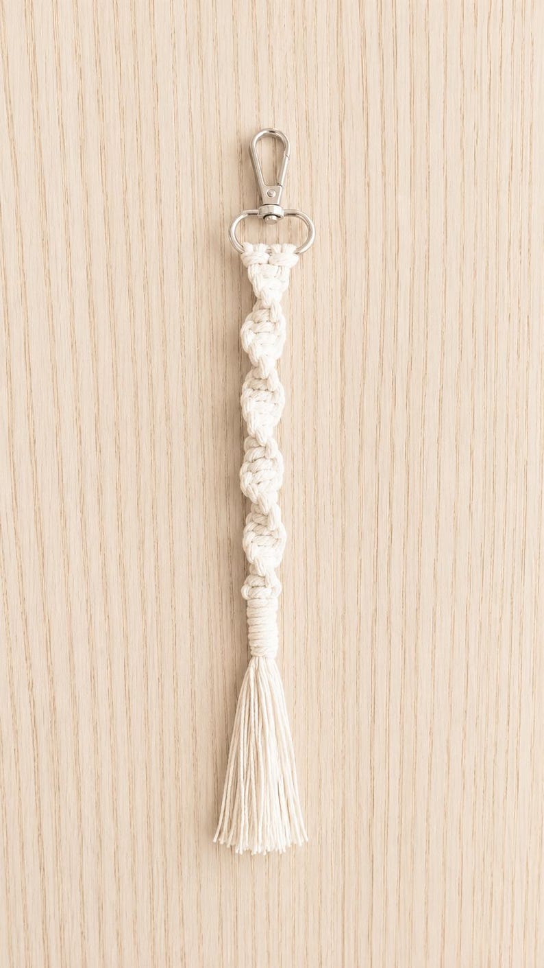 Puede incluir: Un llavero de macram&eacute; color crema con un cierre plateado. El llavero presenta un dise&ntilde;o anudado y una borla en la parte inferior. El fondo es de vetas de madera clara.