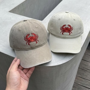 May include: Two baseball caps made of cotton fabric, one in a light beige and the other in a slightly darker tan shade. Each cap features a detailed, embroidered red crab centered on the front panel, with a curved brim and stitched eyelets.