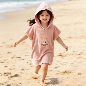 May include: A child wearing a pink hooded beach towel with a bunny and floral embroidery. The towel has the name "Kelly" embroidered on it. The child is running on a sandy beach.