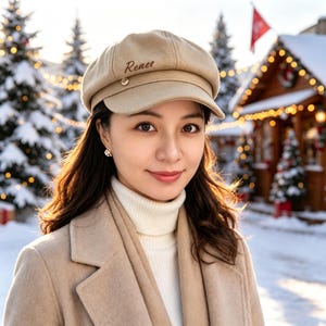 Peut inclure: Une casquette gavroche beige avec le prénom Renee brodé en écriture cursive marron foncé sur le panneau avant, ornée d'un petit bouton métallique sur le côté de la visière, portée avec un pull à col roulé blanc et un manteau en laine beige.