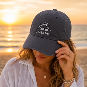 May include: A dark charcoal gray baseball cap featuring a white embroidered graphic of a rising sun over waves with the text Sea La Vie centered below the design. The cap has a curved brim and a structured crown with a button on the top center.