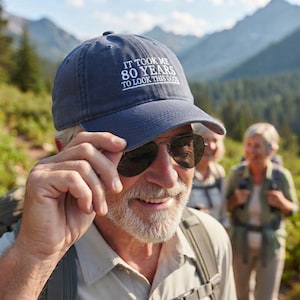 May include: A navy blue baseball cap featuring white embroidered text that reads IT TOOK ME 80 YEARS TO LOOK THIS GOOD. The cap has a structured crown, a curved brim, and a button on top, providing a classic look for outdoor activities and casual wear.